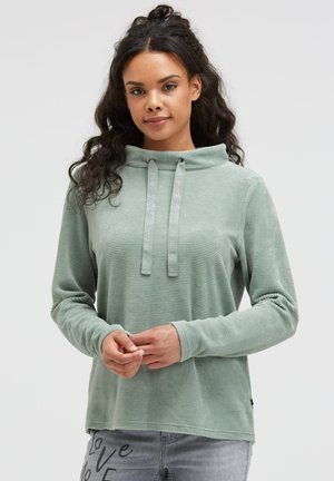 Light green hoodie with ribbed texture, high collar, and drawstrings. Paired with gray jeans featuring a printed accent on the leg.