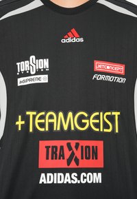 Juoda sportinė marškinėlių su baltais ir pilkais akcentais. Juose yra logotipai, įskaitant raudoną "adidas" ir geltoną "TEAMGEIST".