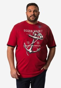 Camiseta roja de manga corta con un gran gráfico de ancla blanca, detalles en negro en las mangas y texto impreso. Fabricada en algodón, de corte casual.