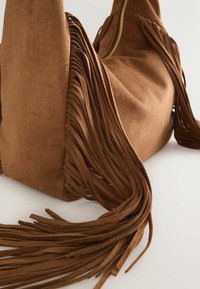 Sac à bandoulière en suède marron avec de longues franges et une fermeture éclair dorée visible sur le côté, posé sur une surface blanche.