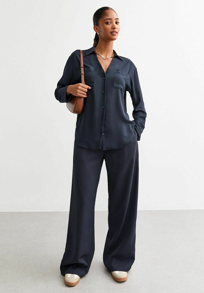 Camicia blu navy con maniche lunghe e tasca sul petto abbinata a pantaloni larghi blu navy e scarpe di colore chiaro. Tessuto dalla texture liscia.