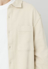 Chemise beige texturée avec une poche, design à boutons, et tissu doux. Caractéristiques : col évasé et motif gaufré subtil.