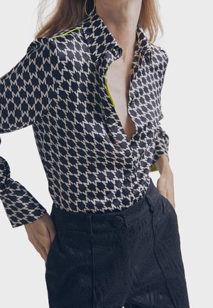 Femme portant un chemisier noir et blanc à motifs en forme de "M" et un pantalon noir texturé taille haute, mains dans les poches, sur un fond blanc.