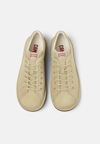 Sneaker in suede beige con punta rotonda, sei occhielli per lacci, collo imbottito e logo rosso "CAMPER" sulla linguetta. Texture liscia e design minimal.
