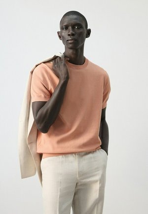 Hombre con camisa de manga corta color durazno y pantalones claros, sosteniendo una chaqueta beige sobre el hombro, de pie frente a un fondo claro y liso.