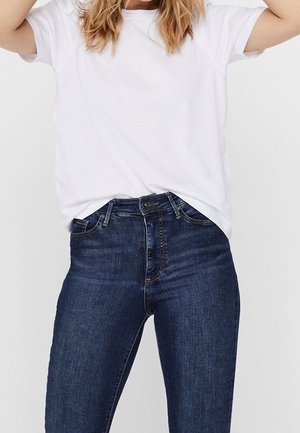 Kobieta ubrana w prostą, białą, krótki rękaw t-shirt włożony w wysokowodne, ciemnoniebieskie dżinsy na neutralnym tle.