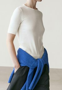 Femme portant un haut crème ajusté à manches courtes avec un pull en tricot bleu noué autour de la taille et un pantalon rayé foncé, mains dans les poches.