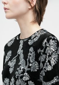 Mulher com cabelo castanho curto, vestindo uma blusa preta adornada com padrões florais de lantejoulas prateadas, voltada para a esquerda, contra um fundo simples.