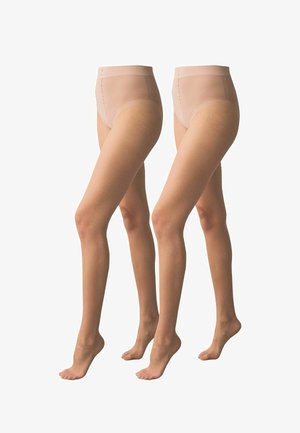 Frau trägt hautfarbene, hochgeschnittene, transparente Strumpfhose und steht barfuß auf einem grauen Boden vor weißem Hintergrund.