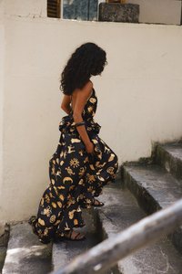 Robe maxi noire avec des motifs dorés imprimés, des détails froncés et des bretelles spaghetti. Le mannequin porte des sandales noires et marche sur des marches en pierre.