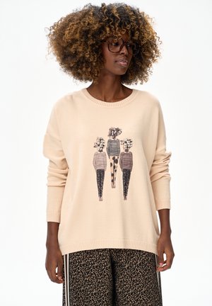 Vrouw met krullend haar, een bril, een beige trui met drie gestileerde figuren met bloemhoofden, en een broek met luipaardprint.