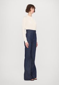 Ljusbeige långärmad topp med knappläppar som är kombinerad med högmidjade mörkblå jeansbyxor med vida ben och kontrasterande sömmar.