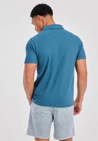 Polo shirt van zacht blauwe stof, korte mouwen, met een gemusterde textuur. Gecombineerd met lichte grijze shorts. Achteraanzicht toont het detail van de kraag.