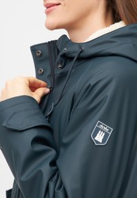 Giacca impermeabile verde scuro con cappuccio foderato in pile, zip frontale, chiusure a scatto e una toppa con logo sulla manica sinistra.