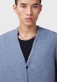 Cardigan di maglia azzurro chiaro, realizzato in morbido materiale, con scollatura a V, chiusura con un singolo bottone e vestibilità comoda sopra un top nero con texture.