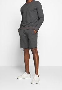 Chemise grise à manches longues avec col et poignets rouges, associée à un short gris avec des poches latérales. Modèle portant des baskets blanches.