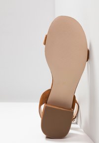 Sandale avec une semelle marron clair et un talon bloc positionnée droite contre un mur blanc d'angle, montrant la semelle texturée et la taille 37.
