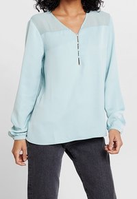 Blouse bleu clair à manches longues, encolure en V et empiècement translucide aux épaules. Présente un détail de patte de boutonnage à trois boutons à l'avant.