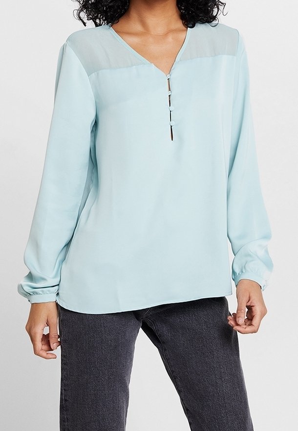 Blouse bleu clair à manches longues, encolure en V et empiècement translucide aux épaules. Présente un détail de patte de boutonnage à trois boutons à l'avant.