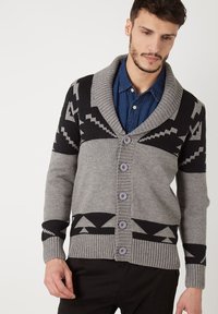 Cárdigan de punto gris con patrones geométricos negros, que presenta un cuello chal y cinco botones en la parte delantera. Puños y dobladillo acanalados.