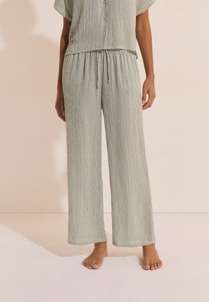 Mujer de pie descalza, vistiendo pantalones anchos de pierna ancha en gris claro con rayas verticales y una blusa a juego con botones.