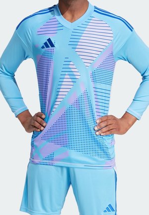 Mann in hellblauem Langarm-Sporttrikot und Shorts mit lila geometrischen und Punktemustern, steht mit den Händen in der Hüfte vor weißem Hintergrund.