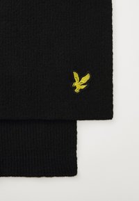 Écharpe noire côtelée avec un logo d'aigle brodé jaune d'un côté. La texture est douce et tricotée, adaptée pour les temps froids.