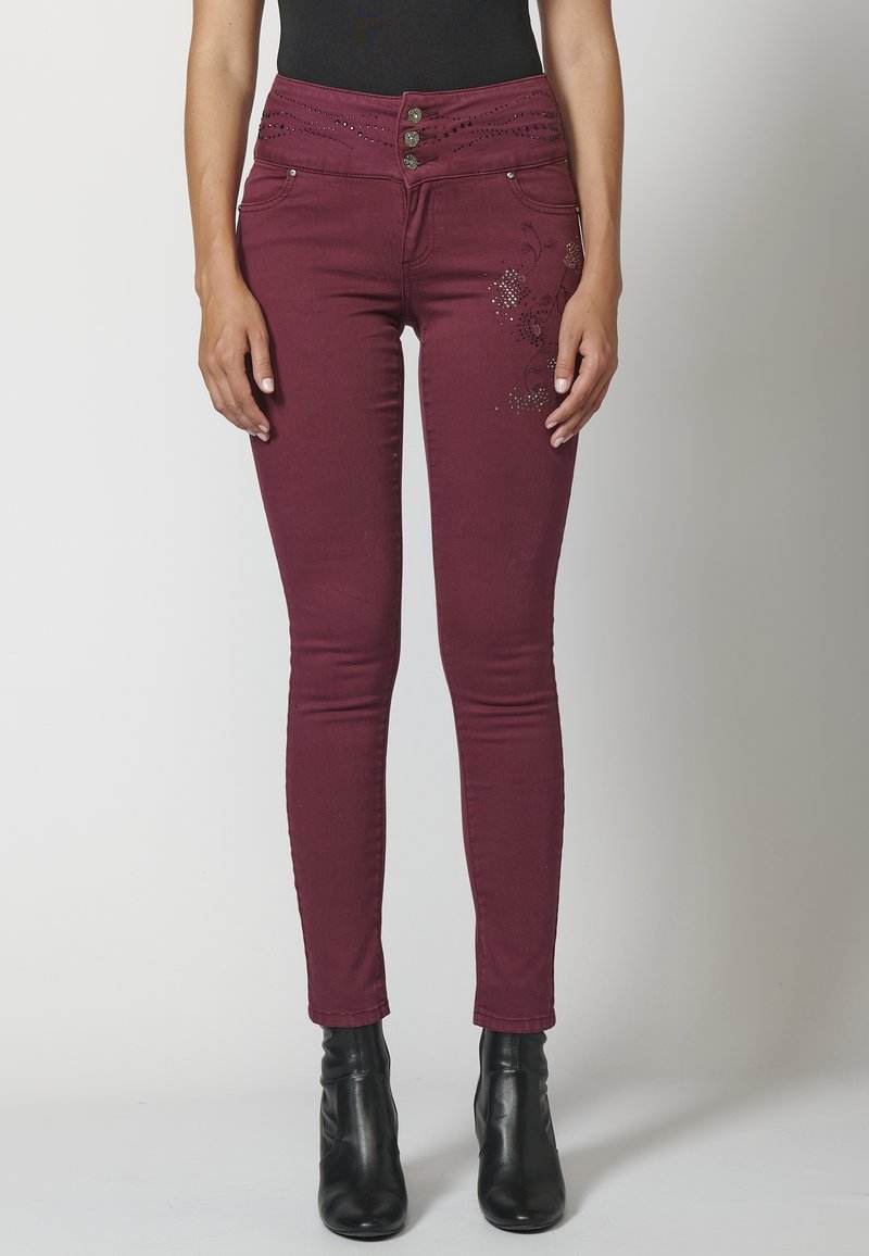 Vaqueros skinny burdeos con cintura alta, que cuentan con un cierre de botón, adornos florales y detalles de strass en la cintura y los lados.