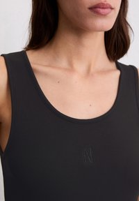 Gros plan sur une femme portant un débardeur noir à côtes avec un petit logo cousu sur la poitrine, montrant le décolleté et une partie du visage.