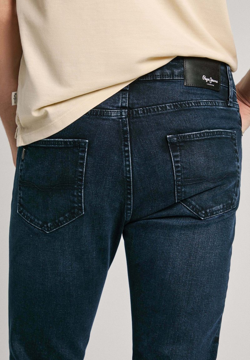 Jeans azul oscuro con una textura ligeramente desgastada, que cuentan con dos bolsillos traseros y un parche de cuero de la marca en la cinturilla.