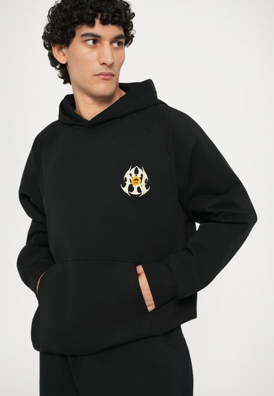 Čierna mikina s klokaním vreckom, s kruhovým vyšitým dizajnom v bielej a žltej farbe na ľavej strane hrude.