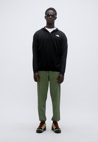 Μαύρο μακρυμάνικο τοπ με λογότυπο, πράσινο παντελόνι και πολύχρωμα παπούτσια. Το μοντέλο φοράει γυαλιά ηλίου. Απλός, εφαρμοστός σχεδιασμός με casual αισθητική.