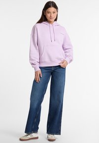 Felpa cappuccio lavanda con tasca frontale, vestibilità rilassata e coulisse. Abbinata a jeans in denim blu a gamba larga e sneakers chiare.