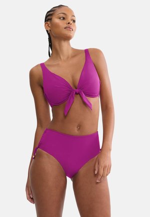 Femeie cu păr împletit, purtând un costum de baie două piese de culoare violet strălucitor, cu top nodat și slipuri cu talie înaltă, pe un fundal simplu.