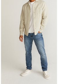 Lyse beige skjorte med knapper over en hvid t-shirt, parret med blå jeans og cremefarvede sneakers; har en brystlomme og en afslappet pasform.