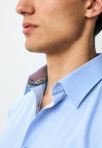 Camicia azzurro chiaro con trama e colletto decorato da motivi a strisce e dettagli floreali. Design con bottoni, che mette in risalto gli accenti interni del colletto.