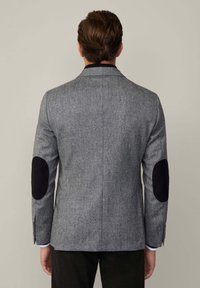 Chaqueta gris de tejido entrelazado con parches de ante negros en los codos, corte entallado, solapas muesca y una abertura en la parte trasera; detalle de tela texturizada.