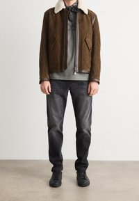 Chaqueta de ante marrón con cuello de borrego, camiseta gris, jeans oscuros y zapatillas negras. La chaqueta tiene cremallera frontal y bolsillos laterales.