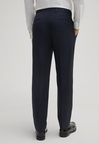 Pantaloni da dress blu navy, vestibilità su misura, tessuto liscio, due tasche posteriori con chiusura a bottone, indossati con una camicia bianca a maniche lunghe e scarpe nere.