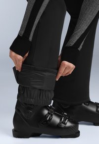 Černá lyžařská bota s nastavovacími přezkami, spárovaná s černými kalhotami s texturovaným pasem a elastickými dolními manžetami pro pevné uchycení.