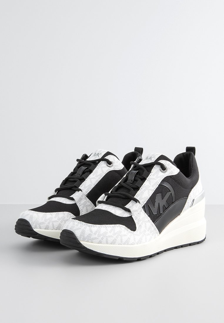 Une paire de baskets compensées noires et blanches avec des lacets noirs et un logo "MK" sur le côté, exposée sur une surface grise.