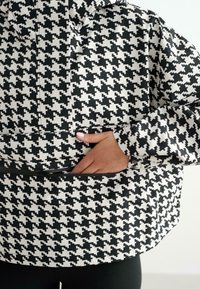 Hand in de voorkeurszak van een zwart-wit houndstooth patroon jas met een rits, gedragen over zwarte broek.