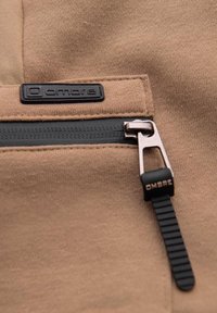 Gros plan d'une poche en tissu beige avec une fermeture éclair noire et une languette noire portant l'inscription "OMBRE" sur un vêtement.