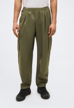 Άνδρας που φοράει χακί παντελόνια με ελαστική μέση και μαύρα παπούτσια πεζοπορίας, στέκεται μπροστά σε λευκό, απλό φόντο.