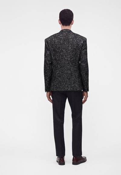 Homme debout, dos à la caméra, portant un blazer texturé noir et blanc, un pantalon noir et des chaussures de ville marron foncé.