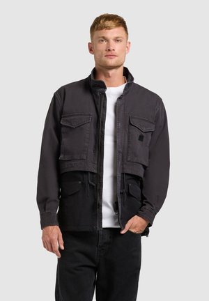 Mann, der frontal eine schwarze kurze Jacke mit zwei Brusttaschen, ein weißes Hemd und schwarze Hosen vor einem einfarbigen grauen Hintergrund trägt.