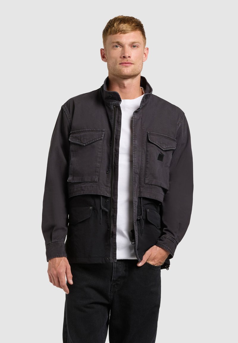 Homme de face portant une veste noire courte avec deux poches poitrine, une chemise blanche et un pantalon noir sur un fond gris uni.