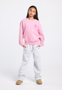 Roze sweatshirt met een ronde halslijn en geribde zoom, gecombineerd met lichtgrijze joggingbroek. De outfit wordt aangevuld met beige instapschoenen.