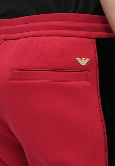 Punaiset verryttelyhousut mustilla samettisilla sivukoristeilla, joissa on takatasku ja pieni kultainen logo vyötäröllä. Sileä tekstuuri, joustava vyötärönauha.