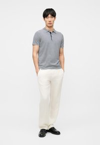 Jeune homme debout, mains dans les poches, portant un polo à manches courtes rayé bleu marine et blanc, un pantalon ample crème et des chaussures noires sur fond blanc.